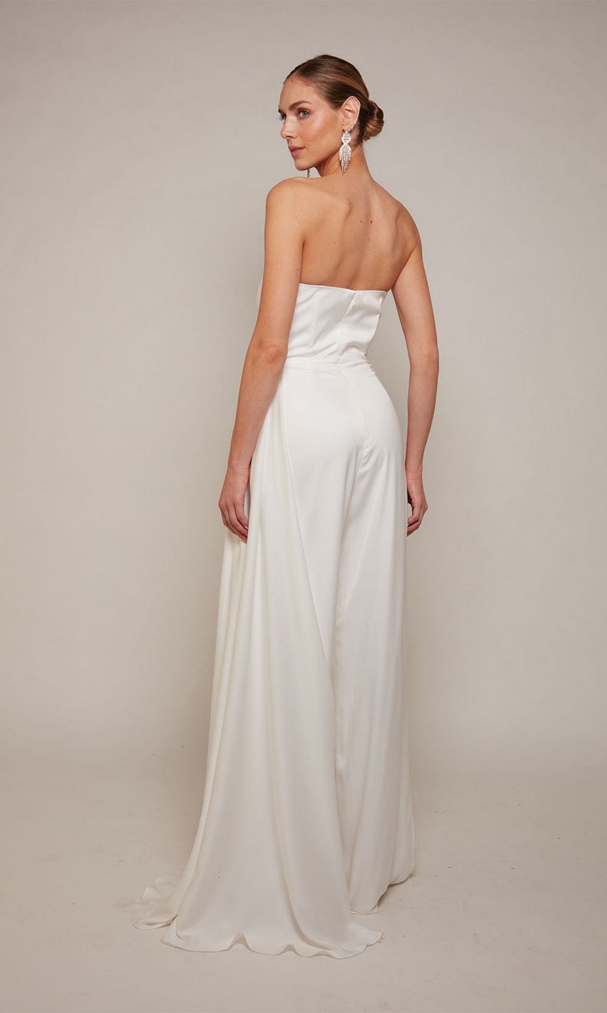 A woman wearing the Alyce 70050 Long Formal Jumpsuit, featuring a strapless fitted bodice and flowing skirt, stands against a plain light background, facing slightly away from the camera.
